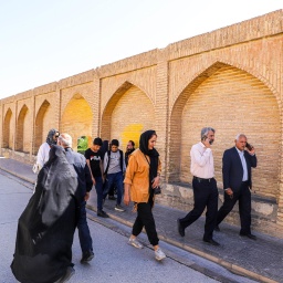 In der iranischen Stadt Isfahan laufen mehrere Passanten auf einer Straße entlang, neben der Straße steht eine Mauer mit Bögen.