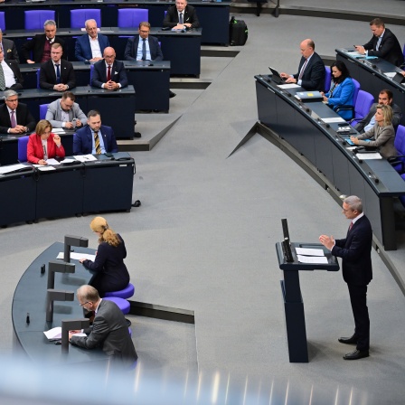 Eine Sitzung im Deutschen Bundestag, dem Parlament der Bundesrepublik Deutschland. In einem großen, modernen Saal sitzen zahlreiche Abgeordnete in mehreren Reihen auf blauen Stühlen, die ordentlich hinter schwarzen Tischen angeordnet sind. 