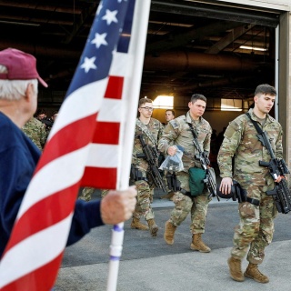 US-Soldaten gehen aus einer Halle zum Flugzeug und dort steht ein Veteran mit der USA-Flagge in der Hand.