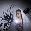 Ein Mädchen sitzt unter der Bettdecke mit einer Taschenlampe und an der Wand sind Schatten.