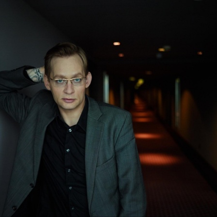 Clemens Meyer lehnt in einem dunklen Hotelflur an einer Wand und schaut in die Kamera. 