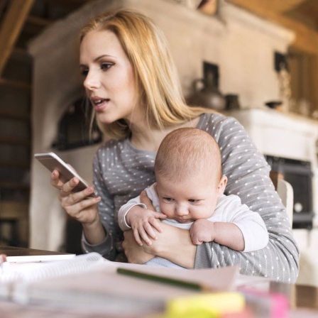 Eine Frau sitzt mit Baby im Arm und Smartphone in der Hand vor einem Notebook
