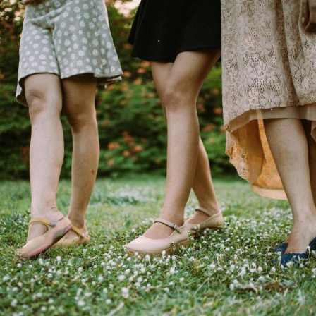 Die nackten Beine von drei Frauen in Röcken und Schuhen sind zu sehen. Sie spazieren über eine Wiese.
