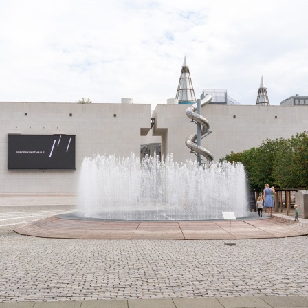 Man sieht die Fassade der Bundeskunsthalle Bonn. Davor ist ein Springbrunnen, auf dessen Rand eine Frau mit Kindern spaziert.