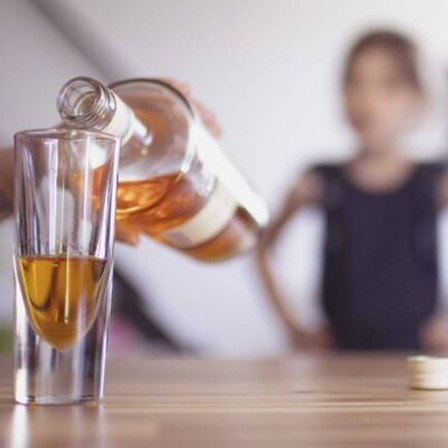 Hand gießt Whisky ins Glas, im Hintergrund ist unscharf ein Kind zu erkennen