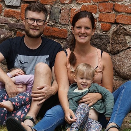 Eine Frau und ein Mann sitzen mit zwei Kindern auf dem Boden vor einer Backsteinmauer.