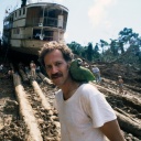 Werner Herzog am Set zu "Fitzcarraldo" mit einem grünen Papageien auf der Schulter und einem auf Baumstämmen transportierten Dampfschiff im Hintergrund.