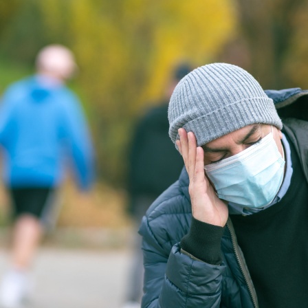 Mann mit Mütze, medizinischer Maske und geschlossenen Augen hält sich den Kopf