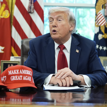 Donald Trump einen dunkelblauen Anzug, ein weißes Hemd und eine rote Krawatte. Auf dem Schreibtisch vor der Person liegen mehrere rote Hüte mit dem Slogan "MAKE AMERICA GREAT AGAIN" in weißen Buchstaben. Außerdem sind einige Dokumente und ein Stift auf dem Schreibtisch zu sehen. Im Hintergrund befinden sich Flaggen und Embleme, darunter eines, das anscheinend mit dem United States Marine Corps zu tun hat.