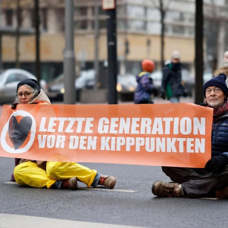 Drei Personen in Winterkleidung sitzen auf der Straße. Die Person links und die Person in der Mitte haben eine Hand flach auf der Straße. Die Person rechts hält gemeinsam mit der Person in der Mitte ein Transparent mit der Aufschrift: "Letzte Generation vor den Kipppunkten".
