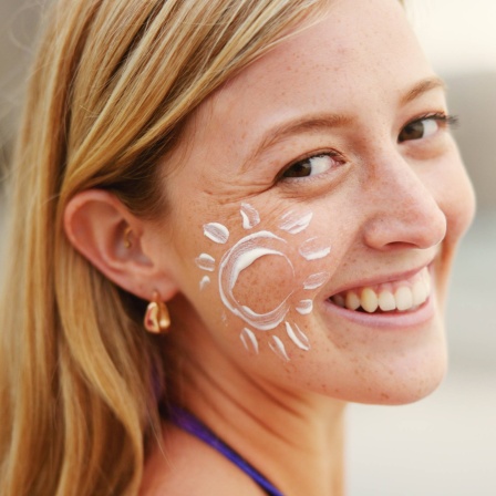 Junge Frau mit Sonnencreme in Form einer Sonne auf der Wange lacht in die Kamera: Die Haut ist unser größtes Organ. Wie bleibt meine Haut gesund und was kann ich dafür tun?