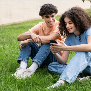Ein lächelndes Paar sitzt im Park auf einer Wiese und schaut gemeinsam in ein Smartphone.