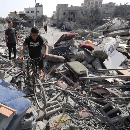 Ein Junge schiebt sein Fahrrad über Trümmer eines Hauses im Gazastreifen, im Hintergrund steht ein Mann.