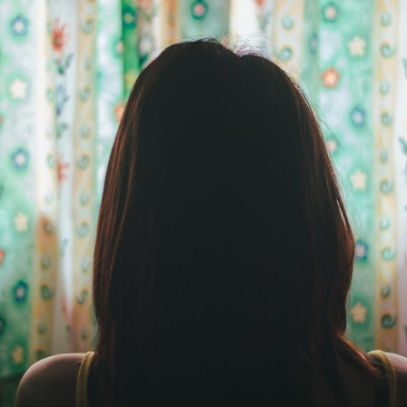 Eine Frau mit langen, braunen Haaren sitzt mit dem Rücken zur Kamera vor einem bunten Vorhang.
