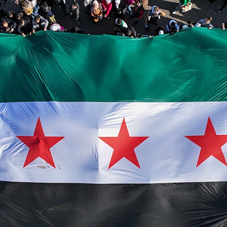 In Damaskus halten Menschen eine große Fahne mit drei roten Sternen in der Mitte, es ist die Fahne der Opposition. Der Blick fällt von oben auf die Flagge und die Menschen (13.12.2024)