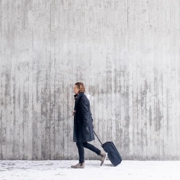 Eine Frau vor einer grauen Wand zieht einen Koffer hinter sich her. 