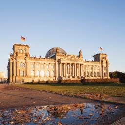 Das Berliner Reichstagsgebäude aus größerer Entfernung fotografiert. Das Gebäude spiegelt sich in einer Wasserpfütze.