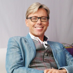 Der Moderator, Sänger, Entertainer und offizielle Schwarzwaldbotschafter Hansy Vogt sitzt mit einem blauen Jeans-Jackett auf einem weißen Sofa und grinst freundlich in die Kamera.