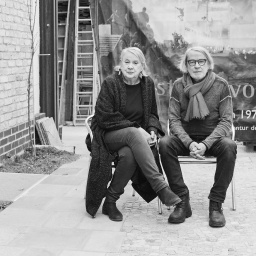 Ute und Werner Mahler sitzen auf zwei Stühlen in einem Innenhof. Sie trägt eine lange schwarze Strickjacke und große Ohrringe. Er trägt einen hellen Schal und eine schwarz umrandete Brille. 