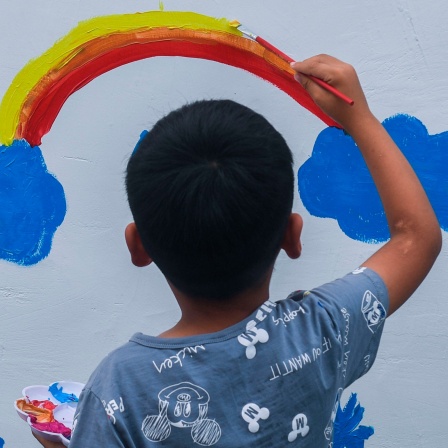 Ein Junge malt mit bunter Farbe einen Regenbogen auf eine Wand.
