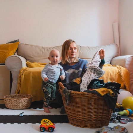 Eine Mutter sitzt im Wohnzimmer neben ihrem Sohn auf dem Fußboden und sortiert Wäsche. 