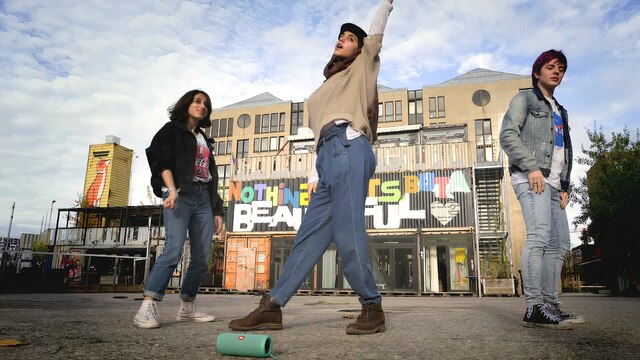 Drei Jugendliche tanzen vor einem Gebäude, an dem der Schriftzug "Nothing lasts but a beautiful heart" zu lesen ist.