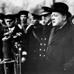 Winston Churchill steht an mehreren Mikrofonen und spricht zu Marine-Soldaten, die im Hintergrund zu sehen sind. 