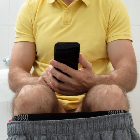 Ein Mann sitzt mit heruntergelassener Hose und Smartphone auf der Toilette.