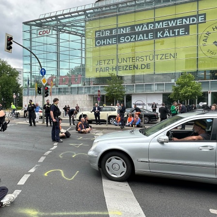 Ein Klima-Aktivist sitzt an einer Straßenkreuzung, neben ihm ein Polizist.
