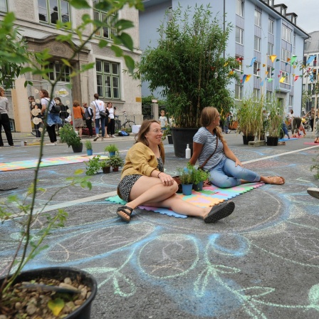 Symbolbild: Menschen sitzen neben Grünpflanzen auf einen bemalten, autofreien Straße, daneben ein Schild mit der Aufschrift "Straßenpicknick - Hock di nieder!"