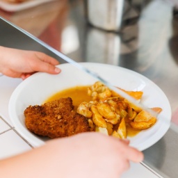 Ein Kind in einer Schulmensa nimmt einen Teller mit paniertem Fleisch und frittierten Kartoffelecken entgegen