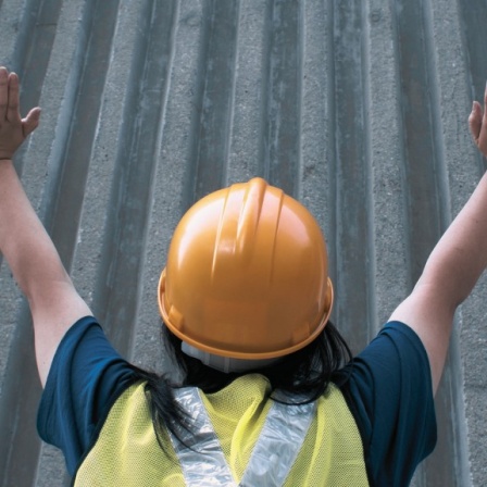 Das Bild oben zeigt eine Frau mit einem gelben Baustellenhelm, die vor einer grauen Betonwand steht. Sie hat die Arme erhoben. 
