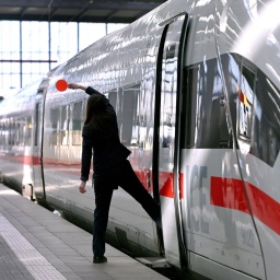 Eine Zugbegleiterin ist von hinten zu sehen, wie sie mit einem Bein auf dem Bahnsteig,  mit dem anderen Bein schon im Zug steht. Sie hält eine Kelle in die Luft.