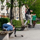 Ein Skateboarder schläft auf einer Parkbank während ein weiterer Skateboard hinter ihm auf einer Parkbank einen Trick aufführt.