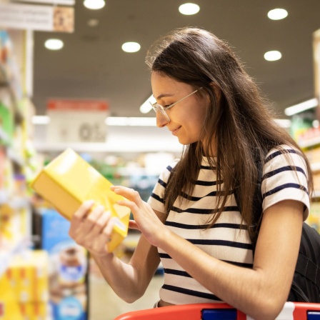 Frau liest im Supermarkt die Informationen auf einer Verpackung: Die Angaben auf Lebensmittelverpackungen sind noch immer irreführend: Sie gehen von einem durchschnittlichen Kalorienbedarf von 2.000 pro Tag aus. Diese &#034;Kalorien-Lüge&#034; hat die Wissenschaft berichtigt.