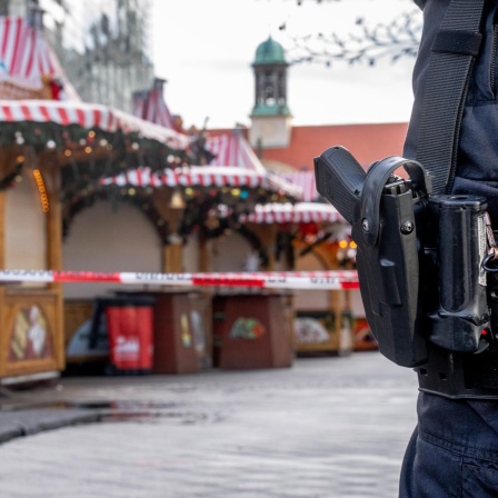 Eine Schusswaffe am Gürtel eines Polizeibeamten, im Hintergrund ist ein Absperrband und der Magdeburger Weihnachtsmarkt zu sehen.