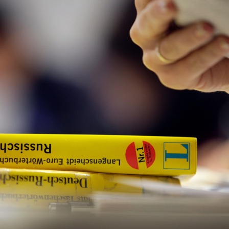Auf einem Tisch bei einem Sprachkurs liegen zwei Wörterbücher für Russisch, während die Hand einer Frau ein Arbeitsblatt hält.