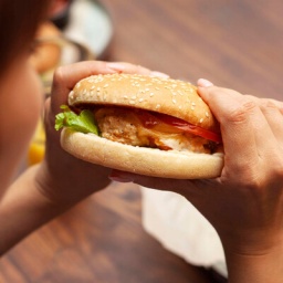 Eine Frau beißt in einen Hamburger, auf dem Tisch steht ein Teller mit Pommes Frites.
