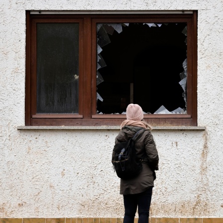 Das Beitragsbild des Dok5 Feature "Wasser bis zum Hals" zeigt eine junge Frau vor einer Hausfassade vor einem zerborstenen Fenster stehen.