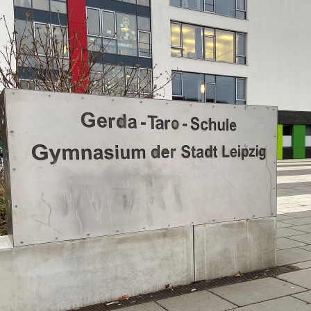Ein Schild an einem Betonblock auf dem der Name einer Schule steht, im Hintergrund sieht man das Schulgebäude