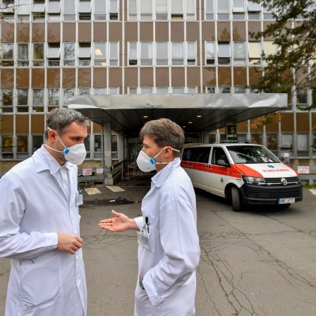 Zwei Personen mit Mundschutz stehen in weißen Kitteln vor einem Krankenhausgebäude