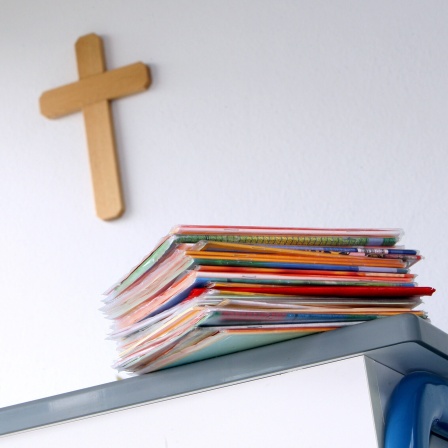 ILLUSTRATION - Ein Kreuz hängt im Klassenzimmer einer Grundschule hinter dem Lehrerpult mit Schulheften.