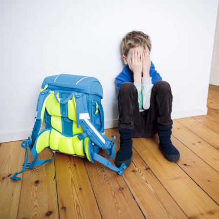 Ein Junge sitzt traurig in seinem Kinderzimmer neben seinem Schulranzen und versteckt sein Gesicht hinter seinen Händen