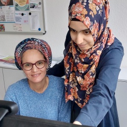 Zwei Frauen sitzen vor einem Computer und sprechen miteinander.