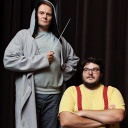 Das Bild zeigt Devid Striesow, der einen Kaputzenmantel trägt und auf einem Podest steht, einen Taktstock hält. Rechts neben ihm steht Axel Ranisch mit verschränkten Armen.