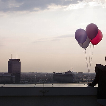 Zwei Personen stehen auf einer Dachterrasse und blicken auf Berlin, die Sonne scheint. Sie halten vier bunte Luftballons in der Hand.