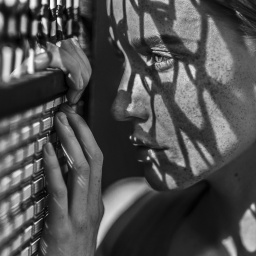 Schwarz-weiß: Eine Frau schaut durch einen engmaschigen Zaun, auf ihr Gesicht fallen die Schatten der Maschen.