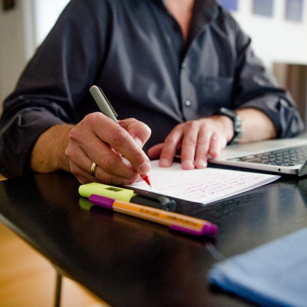Ein Mann sitzt zuhause an einem Esstisch. Er schreibt mit einem Stift auf ein Blatt Papier neben einem Laptop.
