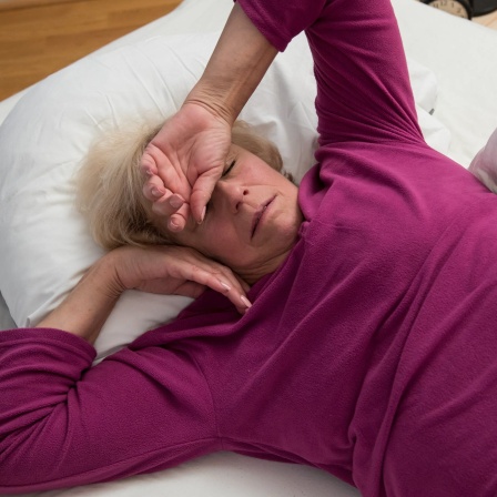 Ältere Frau, aufgedeckt im Bett, fühlt sich unwohl und reibt sich die Stirn: Zu den klassischen Beschwerden in den Wechseljahren gehören, bedingt durch den Östrogenmangel, Hitzewallungen und Schweißausbrüche, aber auch Gelenkbeschwerden. Denn auch Knochen brauchen Östrogen.