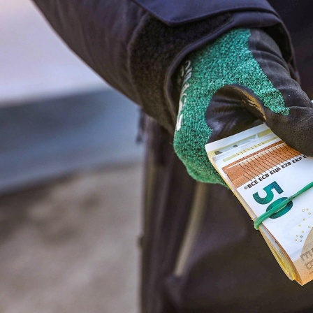Eine Person hält ein Bündel mit Geldscheinen in einer Hand mit Handschuhen.